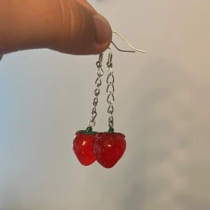 Jordgubbsörhängen med kedja. Handgjorda - Jag gör egna smycken. Supergulliga örhängen med röda jordgubbar i plast som hänger på en silvrig kedja. Perfekt för dig som vill ha en lekfull och färgglad detalj till din look. Krokarna är i metall och jordgubbarna har små gröna blad upptill.