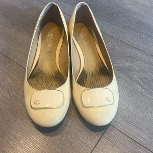 Krämvita ballerinaskor från Lauren Ralph Lauren - Snygga krämvita ballerinaskor från Lauren Ralph Lauren med lackad finish och dekorativ RL-detalj framtill. Skorna har rund tå och platt sula, perfekta för en stilren look. Klassisk och enkel design som passar till mycket. 