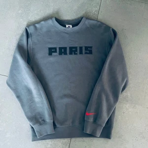 Grå Nike Paris sweatshirt PSG - Snygg grå sweatshirt från Nike x PSG med PARIS broderat på bröstet och röd swoosh på ärmen. Tröjan har rund hals, långa ärmar och mjukt bomullsmaterial. Perfekt för dig som gillar streetwear och fotbollsvibe.