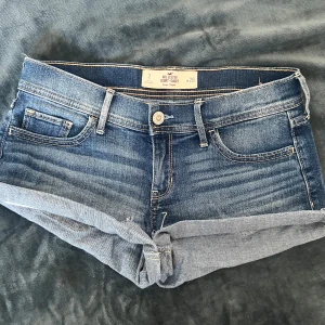 Blå low rise jeansshorts Hollister - Snygga vintage y2k blå jeansshorts från Hollister med låg midja och klassisk femficksdesign. Shortsen har uppvikta benslut och slitna detaljer baktill för en avslappnad vibe. Tillverkade i stretchig denim av bomull och elastan. Perfekta för varma dagar! Short shorts modell. 