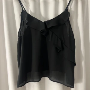 Svart volanglinne från Gina Tricot - Säljer ett svart linne från Gina Tricot med tunna axelband och snygga volangdetaljer framtill. Luftigt och lätt material som ger en skön känsla. Perfekt för dig som gillar en stilren men ändå trendig look.