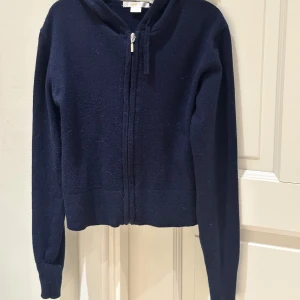 Mörkblå hoodie från Gina Tricot - Mysig mörkblå hoodie från Gina Tricot med dragkedja framtill och huva. Tröjan är i ett stickat material och har långa ärmar. Perfekt för dig som gillar en enkel och stilren look med lite sportig känsla.