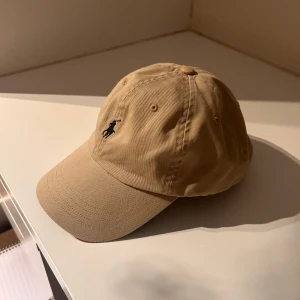 Beige keps från Polo Ralph Lauren - Snygg beige keps från Polo Ralph Lauren med klassisk svart broderad logga framtill och 'POLO' broderat baktill. Justerbar rem i brunt läder baktill för perfekt passform. Tillverkad i bomull, perfekt för en clean och stilren look. Kepsen är ren och fräsch då den blivit tvättad innan försäljning 🫧🧼✨