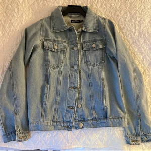 Ljusblå jeansjacka från Boohoo - Snygg ljusblå jeansjacka från Boohoo med klassisk krage, bröstfickor med knappar och raka ärmar. Perfekt för dig som vill ha en tidlös och avslappnad look. Jackan har knäppning framtill och detaljer i silverfärg.