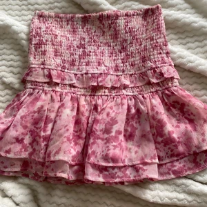 Rosa volangkjol med smock - Supersöt kortkjol i rosa och vitt med smock upptill och volanger nertill. Kjolen har ett blommigt mönster och är perfekt för sommaren💕