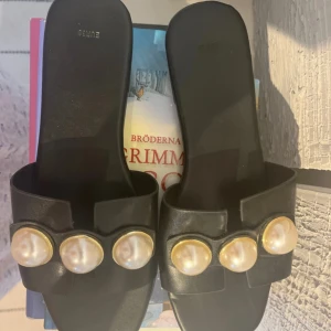 Svarta sandaler med pärldetaljer - Svarta sandaler med platt sula och fyrkantig tå. Över foten sitter ett brett band dekorerat med stora, vita pärlor infattade i guldfärgade ramar. Perfekta för en trendig och chic look under varma dagar. Helt nya 