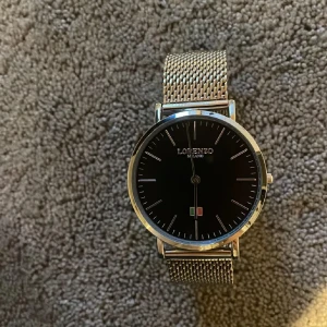 Lorenzo Milano stilren silverklocka - Snygg och minimalistisk klocka från Lorenzo Milano med mesh-armband i silverfärgat metall. Urtavlan är svart med tunna silvermarkeringar och en liten italiensk flagga vid sexan. Passar perfekt för dig som gillar stilren design och klassisk look.