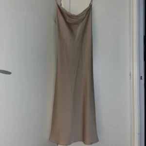 Champagnefärgad lång slipklänning - Säljer en elegant långklänning i champagnefärgad satin med tunna axelband och snygg draperad urringning. Klänningen har en slät, glansig yta och faller snyggt längs kroppen. Perfekt för dig som gillar minimalistisk och stilren look.
