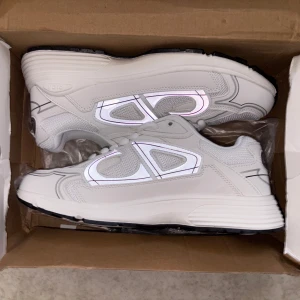 Vita Dior B30 sneakers med reflexdetaljer - Snygga vita Dior B30 sneakers med reflekterande detaljer på sidorna och svart sula. Skorna har meshpaneler för extra andningsförmåga, klassisk snörning och en chunky sula. Perfekta för dig som vill sticka ut med en lyxig och modern look.