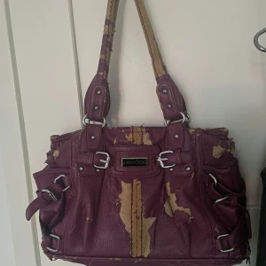 Lila handväska från Jimmy Choo - Säljer en stor och rymlig lila handväska från Jimmy Choo med coola spänndetaljer och silverfärgade metalldelar. Väskan har flera fack och dragkedja, samt breda axelband. Materialet är skinn och väskan har en unik look skriv för fler frågor💜