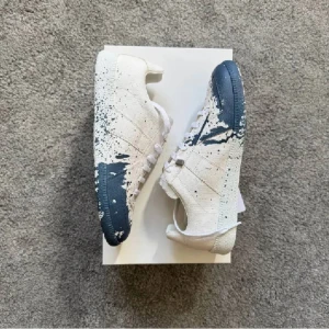 Vitblå sneakers med färgstänk - Unika sneakers i vitt med blå färgstänk över tå och sidor. Snygg design med snörning och platt sula. Perfekta för dig som vill sticka ut med en kreativ och modern look.