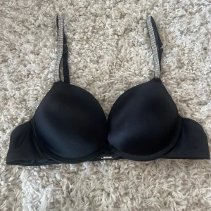 Svart push-up bh med glittriga diamant band - Supersnygg svart push-up bh med glittriga axelband som ger extra bling. Bh:n har släta kupor och en liten metallogo framtill. Perfekt för dig som vill ha något extra under outfiten.
