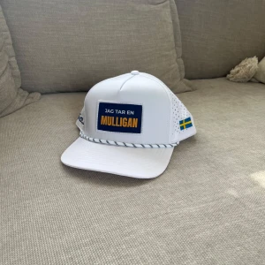 Vit keps Golfio.se Mulligan - Vit golfkeps med blå och gul orange där det står 'Jag tar en Mulligan'. Sidorna är perforerade för extra ventilation och på ena sidan finns en liten svensk flagga. Framtill sitter ett svartvitt snöre längs skärmen. Justerbar baktill för perfekt passform.