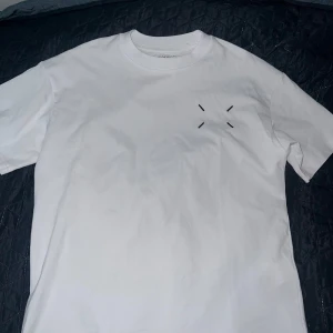 Maison Margiela vit t-shirt med tryck - Vit t-shirt från Maison Margiela med svart grafiskt tryck och logga på ryggen. Klassisk rund hals och korta ärmar. Minimalistisk design framtill med diskreta svarta stygn. Perfekt för dig som gillar stilrena och unika plagg. Storlek M men passar också L