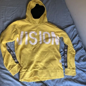 Gul hoodie från WOXO 720 med tryck - Gul hoodie från WOXO 720 med stor vit VISION-tryck på bröstet och svarta band med texten HYPE längs ärmarna. Mjuk insida och huva med svart foder. Perfekt för dig som gillar streetstyle och vill sticka ut.