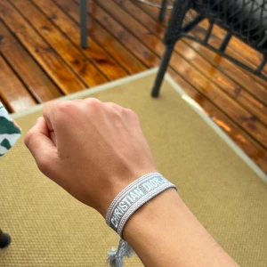 Grått Christian Dior armband - Snyggt vävt armband från Christian Dior i ljusgrått med vit brodyr och tydlig logga. Armbandet har justerbara tofsar i ändarna och ger en clean och trendig vibe till din outfit.