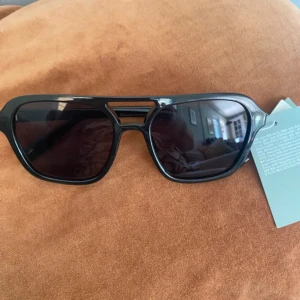 Svarta solglasögon från H&M - Snygga svarta solglasögon från H&M med rektangulära bågar och cool detalj vid näsryggen. Glasen är mörka och ramen är i blank plast, vilket ger en modern och stilren look. 