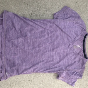 Lila träningst-shirt från SOC - Sportig lila t-shirt från SOC i funktionsmaterial, perfekt för träning. T-shirten har korta ärmar, v-ringning och en diskret reflexdetalj på ryggen. Materialet är lätt och andas, vilket gör den skön att ha på sig under aktiva dagar.