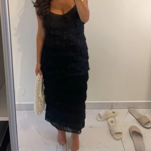 Svart fransklänning med smala axelband - Supersnygg svart klänning med smala axelband och flera lager fransar som ger en cool och festlig look.  Perfekt för dig som vill sticka ut med en trendig och elegant stil. De smala axelbanden är korsade i ryggen. Finns dragkedja vid ryggen som är gömd som gör det enkelt att få på sig klänningen. Klänningen har även en slits nere baktill som är ca 33 cm men jag har sprättat upp den ungefär 5 cm till för att det ska vara lättare att gå i den.