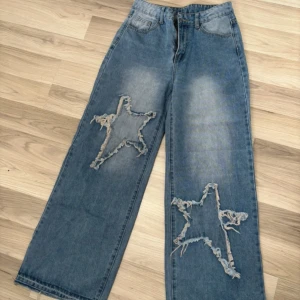Blå wide jeans med stjärn-detaljer - Snygga blå jeans med breda ben och slitna stjärn-detaljer på båda benen framtill. Jeansen har klassisk femficksdesign och en cool, avslappnad vibe. Perfekta för dig som vill sticka ut med ett unikt mönster och loose fit.