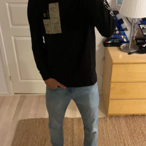 National Geographic svart långärmad tröja - Svart långärmad tröja från National Geographic med tryck av karta och logga på bröstet samt coolt mönster på ryggen. Snygg vit och gul text längs ena ärmen. Skön passform och perfekt för dig som gillar äventyr och streetstyle.