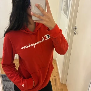 Röd Champion hoodie med logga - Säljer en röd hoodie från Champion. Tröjan har vit Champion-logga över bröstet, huva och en stor magficka. Mjuk och skön bomullsmix, perfekt för chill dagar. Långärmad och klassisk passform. Storlek 152/167 passar dig som bär storlek S/XS. 