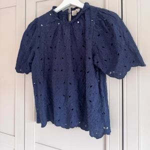 Mörkblå blus med broderade detaljer - Supersöt mörkblå blus med broderade blommor och hålmönster. Puffiga korta ärmar och liten volang vid halsen. 