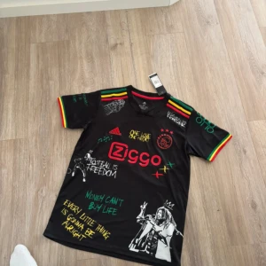 Ajax x Bob Marley svart fotbollströja - Unik svart Ajax fotbollströja från Adidas med Bob Marley-tema. Färgglada ränder i rött, gult och grönt på axlar och ärmslut, tryck med texter och illustrationer, samt Ajax-logga och Ziggo-sponsor. Tillverkad i lätt funktionsmaterial.