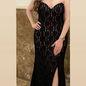 Svart spetsklänning med slits - Supersnygg lång svart klänning med spetsdetaljer och hjärtformad urringning. Klänningen är axelbandslös och har en hög slits framtill som ger en extra touch. Perfekt för dig som vill sticka ut på bal eller fest. Materialet är spets med glittriga inslag. Den är använd en gång!