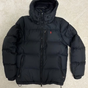 Svart dunjacka Polo Ralph Lauren - Used January–March and still in excellent condition. The jacket shows no signs of wear, and none of the filling has come out from any area. All zippers still work like new! I’m selling it to make room for a new one. Receipt included!  Product details: 	•	Material: 100% polyester (recycled) 	•	Lining: 100% nylon 	•	Outer fabric: 100% polyester 	•	Filling: 75% duck down, 25% duck feathers 	•	Down weight: 650g  