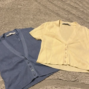 Ribbad croppad topp från Zara - Två ribbade croppade toppar från Zara, en i ljusgul och en i blått. Båda har v-ringning, knappar framtill och kort ärm. Perfekta för en chill och trendig look.