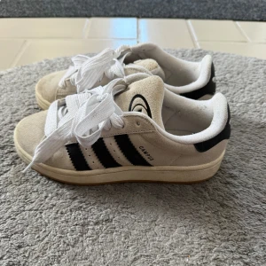 Adidas Campus sneakers i beige mocka - Snygga Adidas Campus sneakers i beige mocka med klassiska svarta ränder på sidorna och vit sula. Skorna har rund tå, platt sula och vita snören. Ikonisk Adidas-logga bak och på plösen. Perfekta för en clean och stilren look.