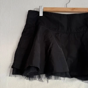 Kort tyllkjol - Svart kort kjol med låg midja och dragkedja i sidan. Lite glansigt tyg i dubbla lager med tyllkant längst ner. Midjemått mätt rakt över ca 39-40 cm. UK size 10 = S eller liten M. Oanvänd, inga defekter. Skriv vid frågor💞