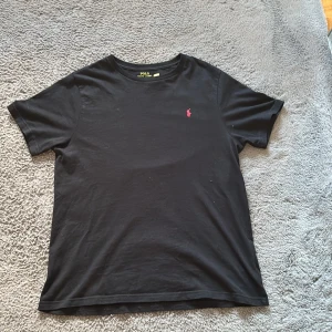 Svart t-shirt från Polo Ralph Lauren - Svart t-shirt från Polo Ralph Lauren i bomull med klassisk passform. Diskret röd broderad logga på bröstet. Enkel och stilren design med rund hals och korta ärmar. Perfekt för en clean och avslappnad look.