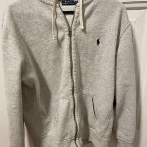 Grå hoodie från Polo Ralph Lauren - Ljusgrå hoodie från Polo Ralph Lauren med dragkedja, huva med snörning och klassisk broderad logga på bröstet. Tröjan har känguruficka framtill och är tillverkad i mjuk bomullsmix. Perfekt för chill dagar eller när du vill ha en clean och enkel look.