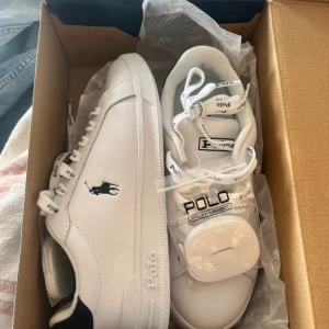 Vita sneakers från Polo Ralph Lauren - Snygga vita sneakers från Polo Ralph Lauren med klassisk logga på sidan och Polo-tryck på snörningen. Skorna har rund tå, platt sula och är tillverkade i syntetmaterial med stilrena detaljer. Perfekta för dig som gillar en clean och sportig look.