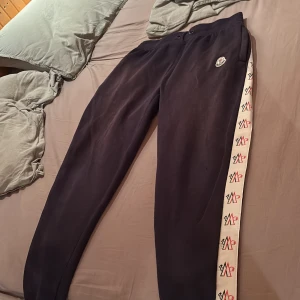 Svarta mjukisbyxor svart tröja från moncler - Svarta mjukisbyxor från Moncler med vit sidoremsa med Moncler-logga i rött och svart längs benen. Byxorna har normal passform och resår i midjan. Perfekta för en chill och sportig look. Även tröjan hör till ett otroligt fint sätt 