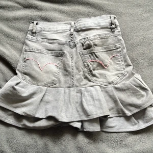 Grå jeanskjol med volang  - Supersnygg grå jeanskjol med bred volangkant och klassiska femficksdetaljer. Baktill finns rosa broderi (som är handsytt) på fickorna som ger en söt touch. Kjolen har bälteshällor och dragkedja framtill. Kjolen är egen gjord och därför lite trådar under volangen. Kjolen i sig är ganska lång och mid waist, kjolen är väldigt stretchig i sig. 