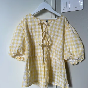 Gul- och vitrutiga blus med puffärm - Supersöt blus i gul- och vitrutigt med puffiga korta ärmar och tre knytningar framtill. Luftig och flowy modell med lätt struktur i tyget, perfekt för varma dagar. Ger en riktigt somrig vibe och är lätt att styla till jeans eller kjol. Aldrig använd!💞