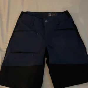 Blå och svarta shorts från Haglöfs - Snygga blå och svarta shorts från Haglöfs i storlek XS. De har flera dragkedjefickor framtill och är perfekta för utomhusaktiviteter. Shortsen har en normal passform och är tillverkade i slitstarkt syntetmaterial.
