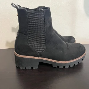 Svarta chelsea boots i mocka - Snygga svarta chelsea boots i mocka med grov sula och platt klack. Resår på sidorna och detaljer i blankt, krokopräglat material baktill. Perfekta för höst och vinter när du vill ha både stil och komfort.
