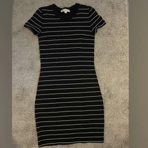 Svart kortklänning Michael Kors - Snygg svart kortklänning från Michael Kors med vita vågiga ränder. Klänningen har rund hals, korta ärmar och en figurnära passform. Perfekt för dig som gillar stilrena och trendiga plagg med en twist.