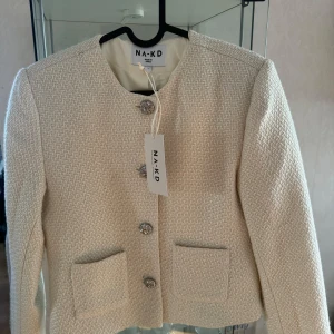 NYTT: Tweed Jacket, stl 32, NAKD, stor i storleken  - Tweed Jacket Den här jackan har ett tweedmaterial och en kort passform. Den har en rundhals och öppna framfickor. Vår jacka har en knappstängning ner längs framsidan. Den här jackan kommer i naturvitt. Komposition: Yttertyg: 55% Akryl, 25% Polyester, 20% Ull (20% Recycled Wool GRS) Foder: 100% Polyester