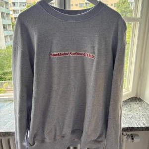 Grå sweatshirt Stockholm Surfboard Club XL - Grå sweatshirt från Stockholm (Surfboard) Club i storlek XL. Tröjan har rund halsringning och tryck på bröstet. Tillverkad i 75% ekologisk bomull och 25% återvunnen bomull. Perfekt för en chill och avslappnad stil.