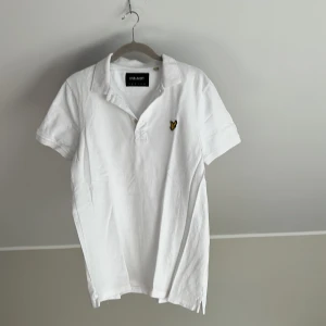 Vit pikétröja från Lyle & Scott  - Vit pikétröja från Lyle & Scott i klassisk modell med korta ärmar och krage. Tröjan har det ikoniska broderade gula örnloggan på bröstet och är tillverkad i bomull. Perfekt för en clean och stilren look.
