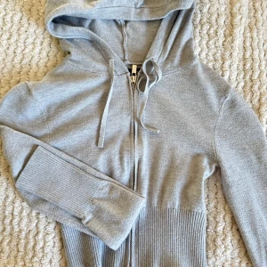 Grå hoodie med dragkedja och huva - Snygg grå hoodie med dragkedja framtill och huva med snörning. Tröjan har långa ärmar och breda ribbade muddar vid ärmslut och midja. Perfekt för en chill och avslappnad stil.
