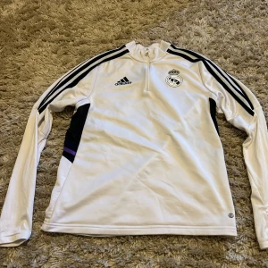 Vit långärmad Real Madrid-tröja Adidas - Snygg vit långärmad fotbollströja från Adidas med Real Madrid-logga på bröstet. Tröjan har svarta ränder längs ärmarna, svart och lila detalj på sidorna samt halv dragkedja vid halsen. Tillverkad i polyester, perfekt för träning eller match.