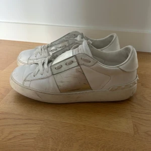 Valentino Garavani Open sneakers vit/silver - Snygga Valentino Garavani Open sneakers i vitt skinn med en metallic silverdetalj över mitten. Klassisk låg modell med snörning och ikoniska nitar baktill på sulan. Perfekta för dig som gillar lyxiga och trendiga sneakers med unik touch.