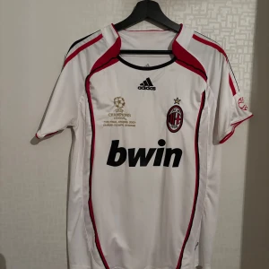 AC Milan Kaka Champions League 2007 tröja - AC Milan bortatröja från Champions League-finalen 2007 med Kaka och nummer 22 på ryggen. Vit med röda och svarta detaljer, broderat klubbmärke och Champions League-emblem. Tillverkad i polyester, klassisk Adidas-design och Bwin som sponsor framtill. Knappt använd 