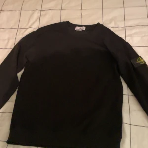 Svart Stone Island tröja - Svart långärmad tröja från Stone Island med den klassiska loggan på vänster ärm. Enkel och stilren design, perfekt för dig som gillar streetwear. Tröjan har rund hals och är gjord i mjukt material för skön känsla.